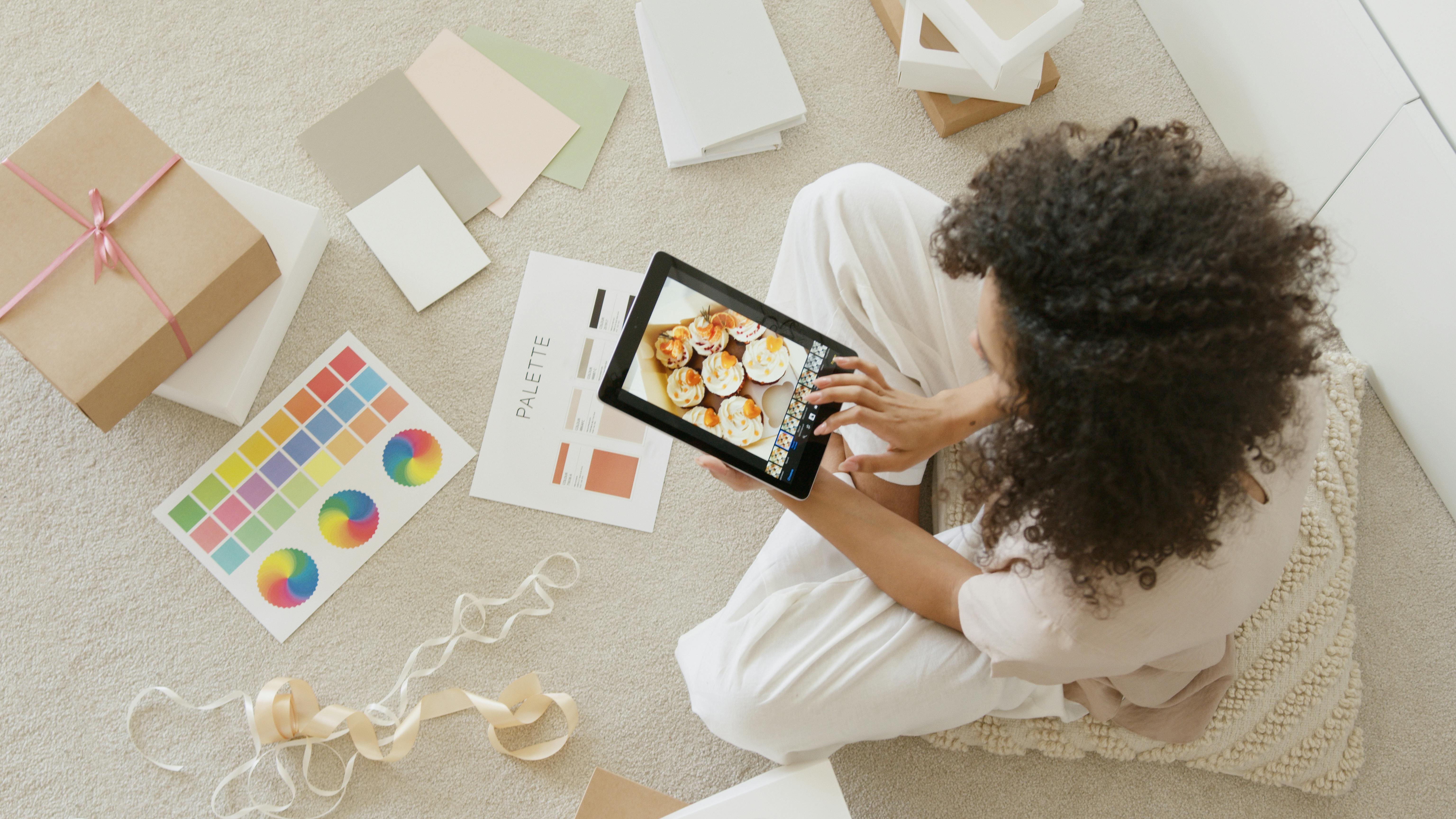 Free Woman using tablet surrounded by design tools and gift box, showcasing creativity and online work. Stock Photo